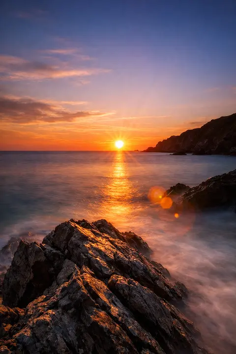 Photoreal sunset wallpaper with a rocky headland, lens flare and long exposure smooth water